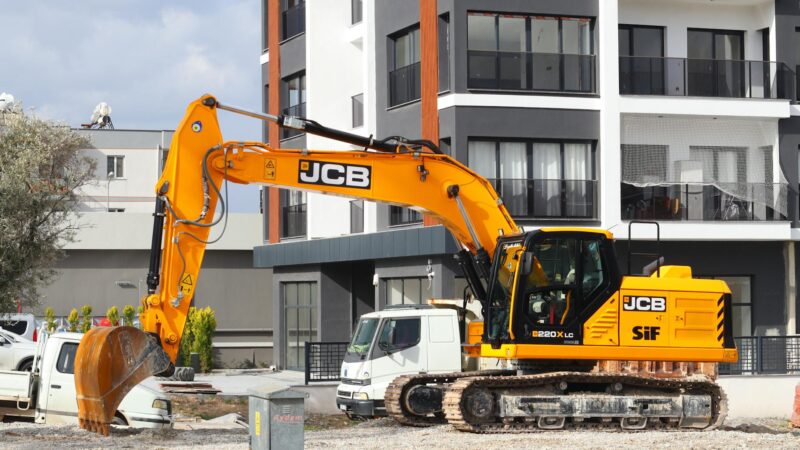 Modern binaların önünde, bir şehir inşaat alanında çalışan sarı bir ekskavatör.